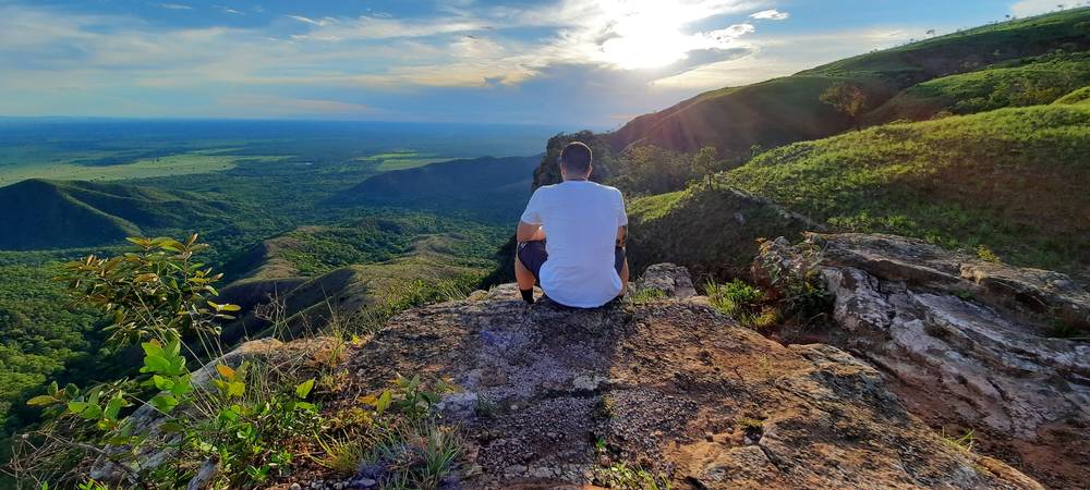 Thiago sentado na beira de um mirante, de costas, olhando o vale verde da chapada
