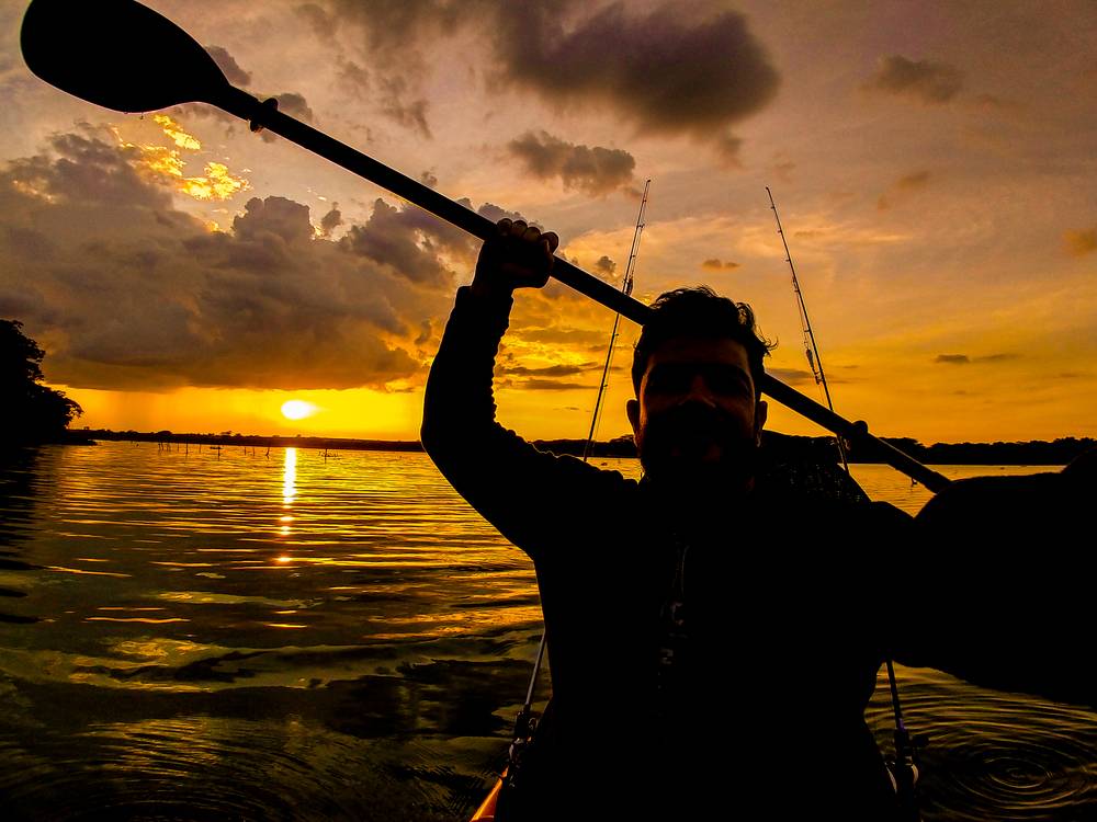 Silhueta de Thiago erguendo o remo em um caiaque no Lago das Garças, ao pôr do sol