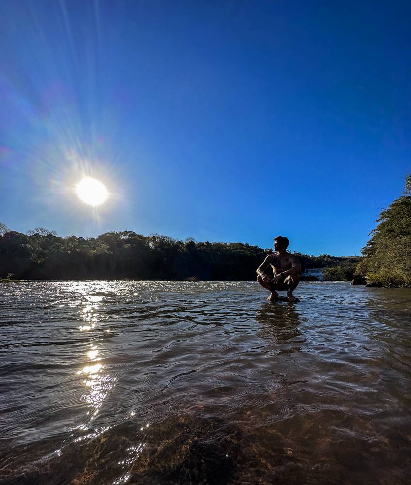 Thiago agachado sobre uma pedra no meio de um rio, sob o sol